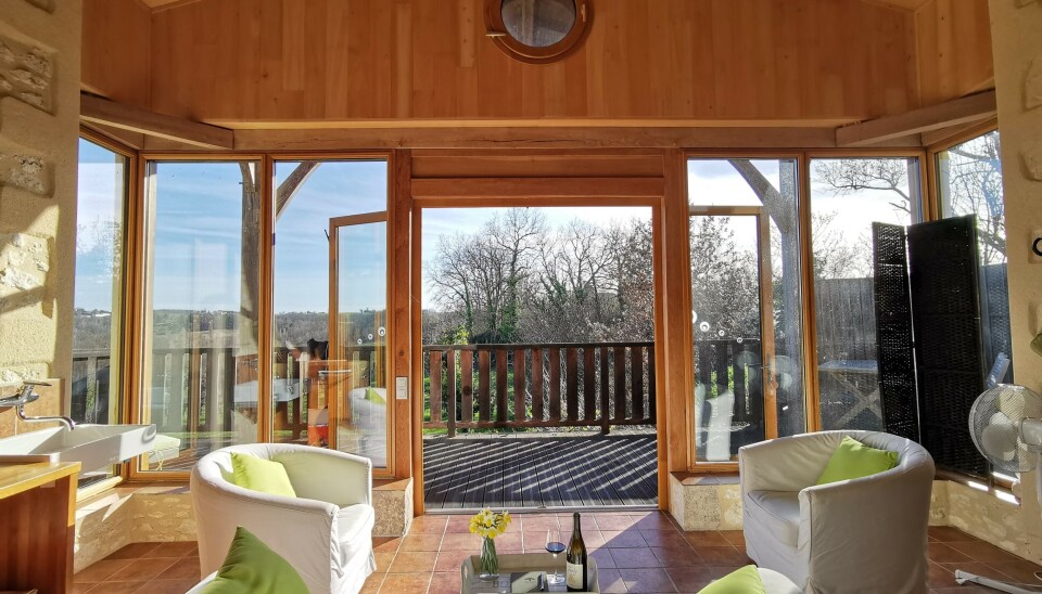 Salon en bois ensoleillé avec fauteuils face aux portes ouvertes sur une terrasse et des arbres
