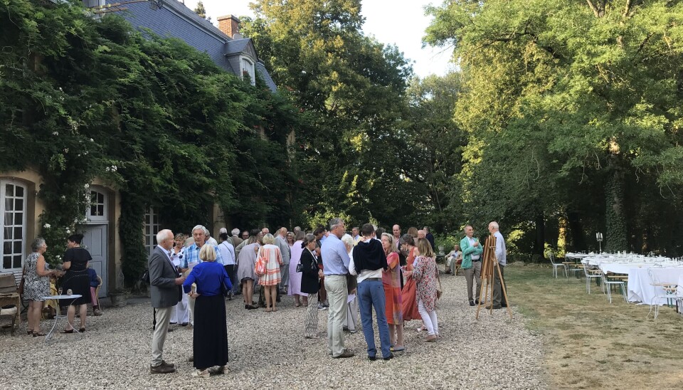 Des gens debout et discutant dans une cour en gravier à côté d’arbres et de chaises blanches lors d’un rassemblement en plein air.