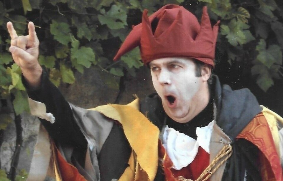 Homme en costume de style médiéval gesticulant avec la main levée devant un feuillage vert.