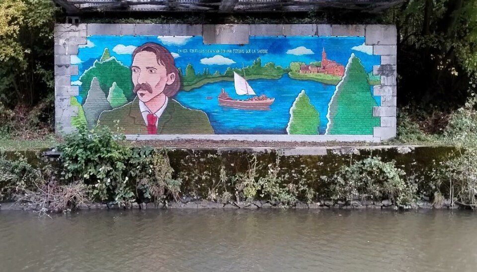 Peinture murale colorée sur un mur au bord d'une rivière, montrant un portrait, des bateaux et des images de paysages.