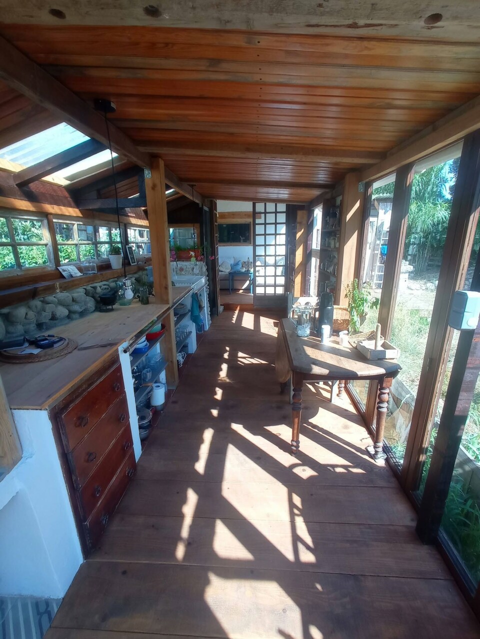 Véranda rustique en bois ensoleillée avec fenêtres, plantes, table et équipements de cuisine.